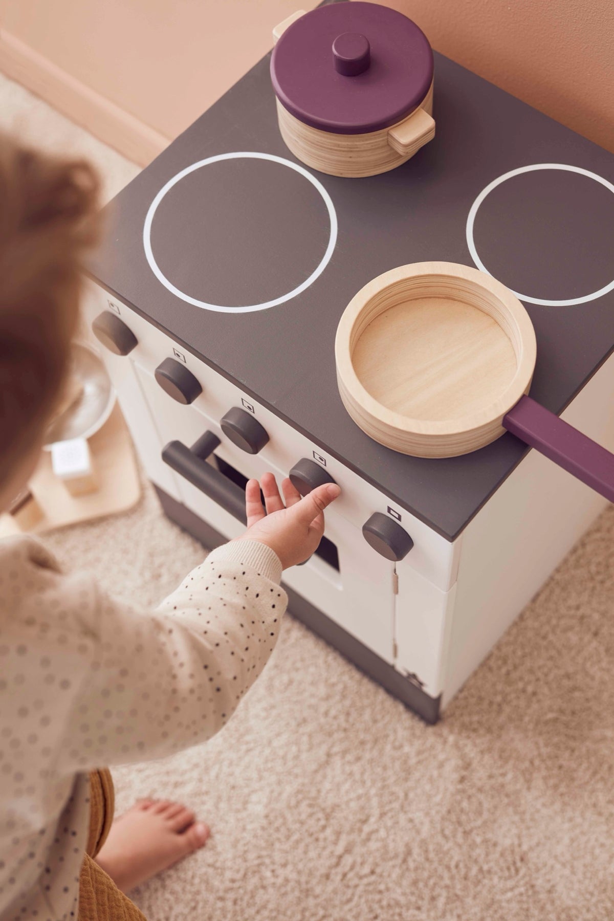 Child turning the knobs of the white wooden toy stove with wooden pots on the hob